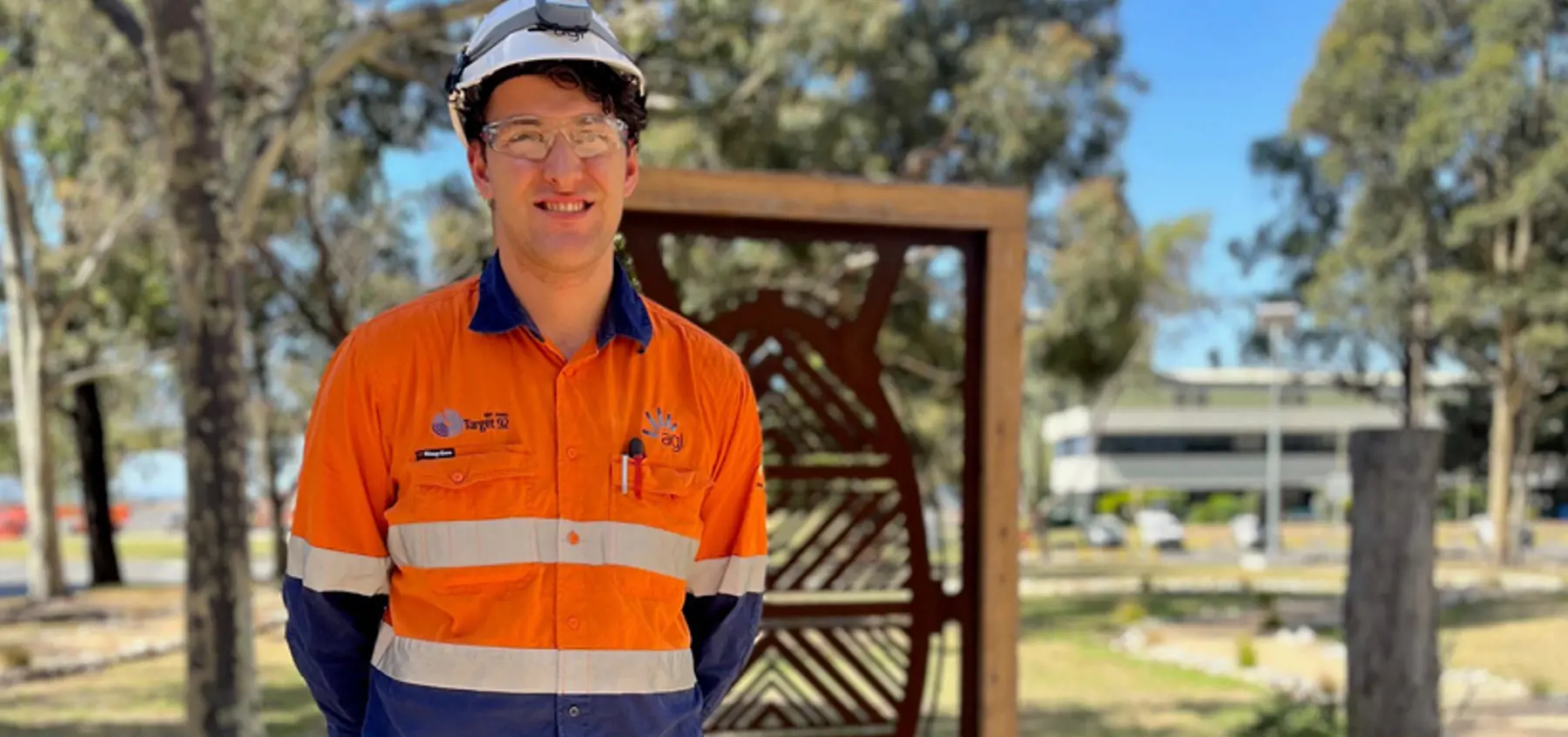 Smiling Federation University co-op placement student Simon Lea on-site at AGL Loy Yang, dressed in hi-vis and hard hat, with trees, buildings and carved wooden panel in background.