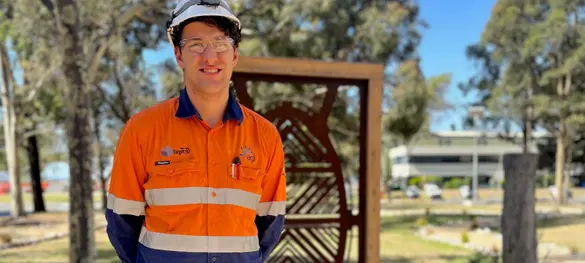 Smiling Federation University co-op placement student Simon Lea on-site at AGL Loy Yang, dressed in hi-vis and hard hat, with trees, buildings and carved wooden panel in background.