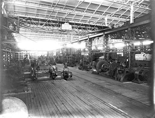 "Black-and-white photograph of the turning and fitting shop at Ballarat North Workshops, showing rows of large industrial lathes and machinery, with workers operating equipment in a spacious, high-ceilinged workshop." (From Turning and fitting shop at Ballarat North Workshops, Museum Victoria. Wikimedia Commons. Accessed Jan. 21, 2025. https://commons.wikimedia.org/wiki/File:Turning_and_fitting_shop.jpg. Accessed Sept. 1, 2025.)
