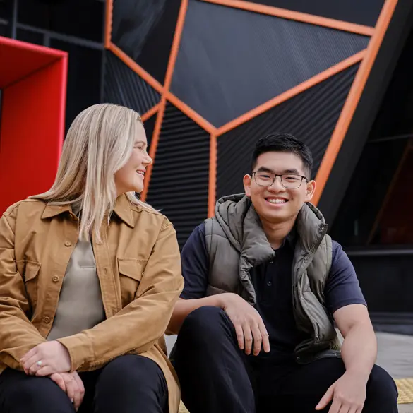 Photo of two smiling people sitting outside in front of a modern building.