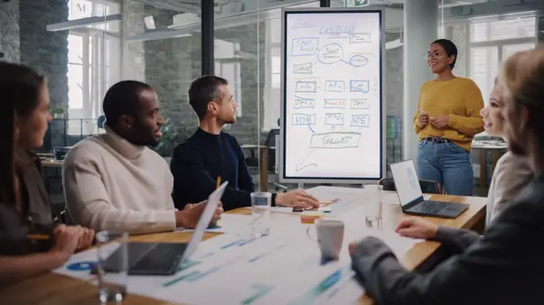 A group of people sitting in a meeting room. A young person is presenting to the group using a whiteboard.