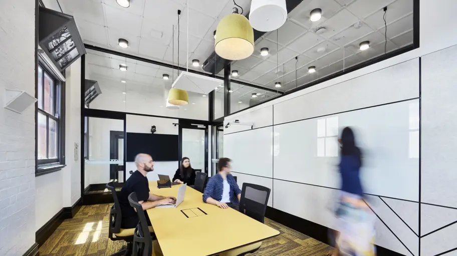 Photo of classroom setting with three people sitting at a big desk and one person near a whiteboard.