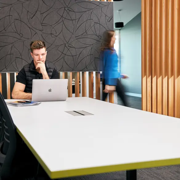 Photo of a young man sitting at a desk using a computer. A blurred woman is leaving the room through a door to the right.