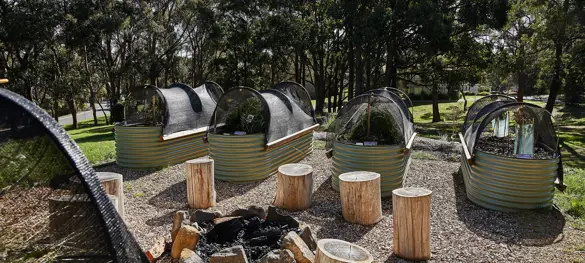 Photo of Wayn-Gurr Derrk Community Gardens showing a central campfire circled by log seating. On either side of the campfire are rows of raised planter beds with shades pinned in an arch over the top and native plants growing within them.
